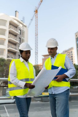 Güvenlik donanımlı iki farklı mühendis inşaat alanının planlarını gözden geçiriyorlar ve arka planda vinç var.