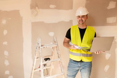 İnşaat sırasında kameraya gülümseyen erkek müteahhit ya da mimar dizüstü bilgisayar ve plan tutuyor.