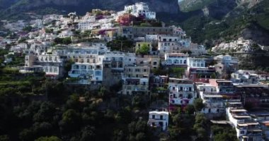 Napoli, İtalya 'daki Positano hava manzarasının yanından geçiyor. Amalfi Sahili boyunca renkli ve beyaz bir kasaba, hareket eden ve yüzen teknelerle beyaz plajı ve mavi okyanusu yüksek bir manzara. Şehrin üzerinde uçuyor.