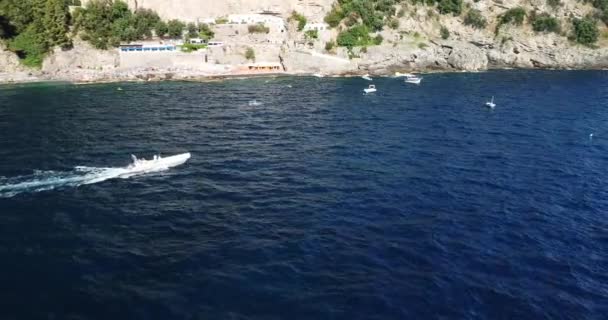 セーリングボートはカメラを通過します 地中海でボートやヨットをセーリング 晴れた夏の日は ビーチの近くを飛んでいます 人々は太陽を楽しんでいる 崖側のビーチ オレンジの太陽の日陰の傘 — ストック動画