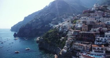 Kayalık kayalıklar, mavi sular, yüzen tekneler, yatlar. Napoli kıyıları, Amalfi, İtalya. Lüks seyahat, tatil, hava aracı görüntüsü. Dağların tepesinde tatil köyleri. İnsanlar plajda güneşleniyor. Yeşiller. 