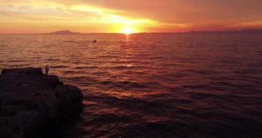 Güneşin doğuşuyla uçurumun kenarında balık tutan yalnız bir balıkçı. Hava aracı görüntüleri, gün batımında balık tutmak için olta. Arka planda dağlar ve yat var. Gün batımının havadan görünüşü. Güneş batıyor.