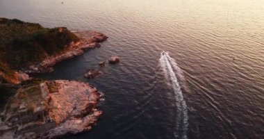  Kayalık kayalıkların yanında gün batımına yakın denizde giden lüks bir yelkenli. Kayalık kayalıklar, mavi sular, yüzen tekneler. Yatçılığın insansız hava aracı görüntüleri, Hayalci Kayalıklar plajı. Dalgalar su sıçratıyor. Tekne gezisi. . 