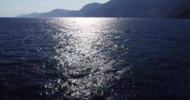 Aerial drone view of the Tyrrhenian sea coast, Positano, Italy. Flying over the Ocean during golden hour. Coastal scene, rocky cliffs, blue water, floating boats, greenery. Waves and boats foaming. 