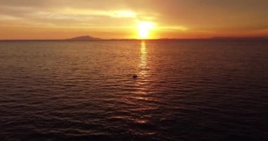 Colorful sunset above the sea surface with sail boat, sun goes down, blue and orange clouds flow in sky. Small yacht boat sailing in calm open sea at sunset. Floating on Sea Surface Golden Sky. 