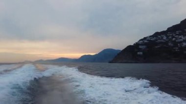 View of the wake water trace behind a ferry at sea at sunrise, sunset. Passing by Positano Mountains, Italy. Wide wake trail from boat back view. Land visible in the background. with sunset sunrise.