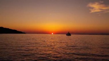 Colorful sunset above the sea surface with sail boat, sun goes down, blue and orange clouds flow in sky. Small yacht boat sailing in calm open sea at sunset. Floating on Sea Surface Golden Sky. 