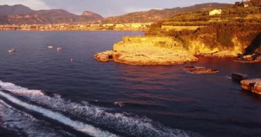 Aerial drone tracking video of luxury yacht cruising in deep blue ocean mediterranean sea next to a rocky coastline. Sunset colors on landscape. Clear beach visited by luxury yachts and sail boats.