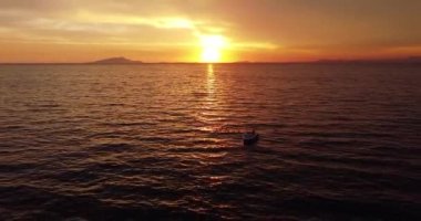 Colorful sunset above the sea surface with sail boat, sun goes down, blue and orange clouds flow in sky. Small yacht boat sailing in calm open sea at sunset. Floating on Sea Surface Golden Sky. 