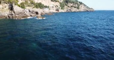 Aerial drone tracking video of luxury yacht cruising in deep blue ocean mediterranean sea next to a rocky coastline. A hotel inside the mountain. Clear beach visited by luxury yachts and sail boats.