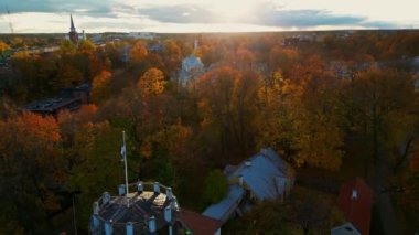 Sunset over city with few clouds in the sky and orange tones. Estonia flag waving in the air with sunset. Early morning sunrise. Colorful autumn trees with golden hour. City and roofs Tartu, Tallinn. 