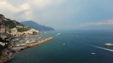 Amalfi sahili hava aracı görüntüsü, eski Amalfi kasabası boyunca uçuyor, deniz kıyısında bir sahil limanı, rıhtım ve botları geçiyor. Postitano, İtalya. Güzel dağ varış yeri. Bulutlu bir gün.