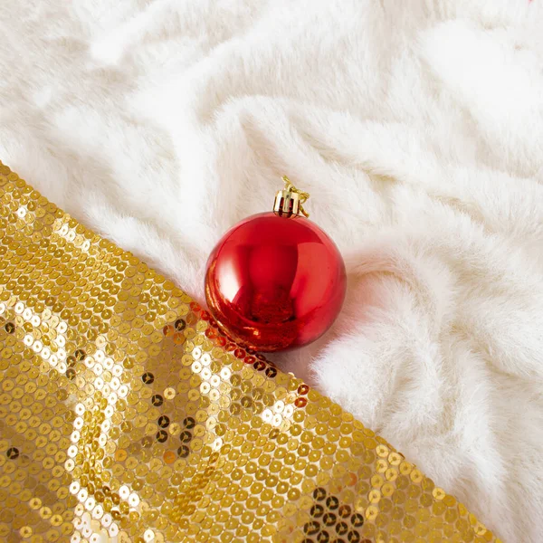 White fur and golden sparkling background with Christmas red bauble. Christmas or New year party concept. Glamour celebation composition.