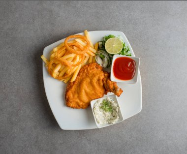 Domates soslu tavuk şnitzel, patates kızartması, limon ve salata. Arka planda gri arkaplan manzaralı Arap yemekleri servis ediliyor.