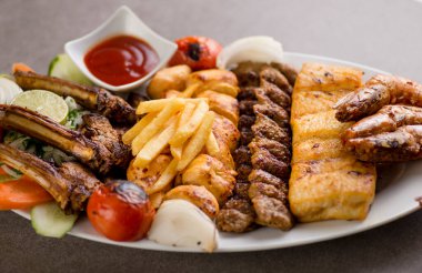 İran usulü ızgara tabağı, tikka boti, kuzu pirzolası, et kebabı, balık, patates kızartması ve domates sosu gri arka planda servis edilir.