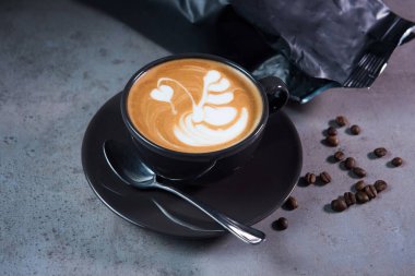 Kahve fincanında servis edilen sıcak kafe Latte sanatı, gri arka planda kaşık ve fasulye manzaralı.