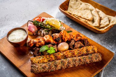 Izgara şiş tawook, tavuk kebabı, sığır kebabı, tikka boti, reshmi ve malai pita ekmeği, nan, tandır roti, raita, sos ve salata, domates soğanı ve limonu ahşap panoda servis edilir.