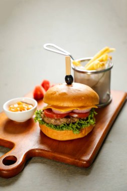 Klasik Cajun Biftek Burger 'i; ahşap tahtada servis edilen peynir dilimi, domates, soğan ve marul yaprağı ile soslu patates kızartması kovası ve aperatif fast food' un gri arka plan manzarası içerir.