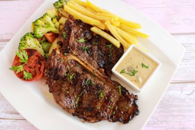 Patates kızartması, mayonez sosu, domates, brokoli ve salatalık salatası ile T-Bone Bifteği, fast food baharatlarının üst görüntüsünde, izole edilmiş tabakta servis edilir.