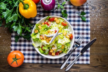 Lahana, domates, kirazlı domates, kızarmış tavuk, soğan, biber, kişniş, bıçak ve çatal peçetede servis edilen tabakta servis edilen peçeteyle servis edilen sağlıklı İtalyan yemeklerini ahşap masada.