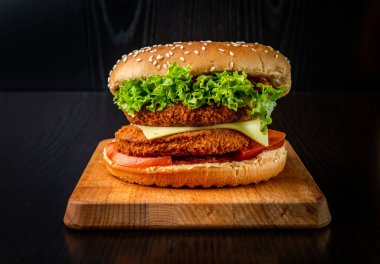 Duble Tavuk Burger peynirli, marullu ve domatesli tahtadan yapılmış, arka planda fast food manzaralı.