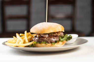 Kızartmalı ve mayonezli klasik sığır burger, beyaz masada ve Amerikan yemeklerinin arka planında izole edilmiş tabakta servis edilir.
