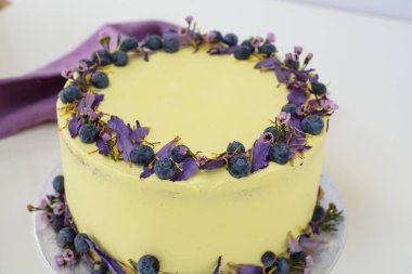 Çiçek desenli limonlu yaban mersinli kek, yaban mersinli ve çiçekli açık sarı bir krema ve peçete köşeli, kutlama için mükemmel yuvarlak pasta manzaralı bir süsleme içerir.