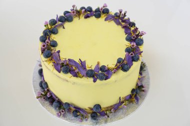 Çiçek desenli limonlu yaban mersinli kek, yaban mersinli ve çiçekli açık sarı bir krema ve kutlama için mükemmel yuvarlak, zarif bir pasta manzarası içerir.