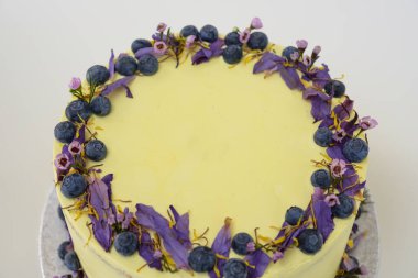 Çiçek desenli limonlu yaban mersinli kek, yaban mersinli ve çiçekli açık sarı bir krema ve kutlamalar için mükemmel yuvarlak, zarif bir pasta manzarası içeriyor.