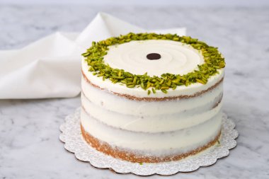 Zarif Pistachio Pastası. Görünür pasta katmanları ve kenarlarında doğranmış şam fıstığı ile süslenmiş pürüzsüz beyaz bir krema. Arka planda gri peçeteyle lezzetli tatlı manzarası.