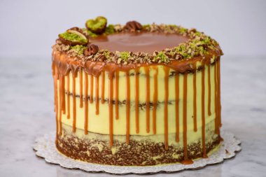 Fıstıklı Karamelli Drip Pastası 'nda zengin karamel serpiştirilmiş, üzerinde şam fıstığı ve fındıklı fındıklı gri arka planda pişmiş tatlı manzarası olan nemli pasta katmanları var.