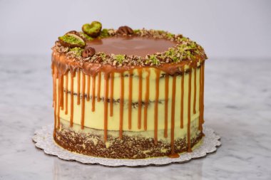 Fıstıklı Karamelli Drip Pastası 'nda bol karamel serpiştirilmiş, üzerinde şam fıstığı ve fındıklı fındıklı gri arka planda pişmiş tatlı kafeteryası manzarası olan nemli pasta katmanları var.