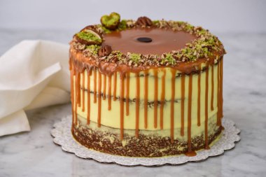 Fıstıklı Karamelli Drip Pastası 'nda bol karamel serpiştirilmiş, üzeri şam fıstıklı ve fındıklı ve peçeteli gri arka planda pişmiş tatlı manzaralı katmanlar var.