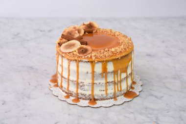 Zarif Karamel İncirli Drip Pastası 'nda zengin karamel serpiştirilmiş, üzerinde kurutulmuş incir dilimleri ve gri arka planda izole edilmiş fındıklar ve fırınlanmış tatlı manzarası olan nemli pasta katmanları bulunur.