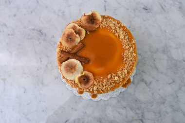 Zarif Karamel İncirli Drip Pastası 'nda zengin karamel katmanları serpiştirilmiş, üzerinde kurutulmuş incir dilimleri ve gri arka planda izole edilmiş fındıklar var.