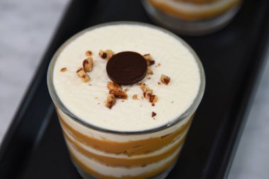 Klasik Tiramisu Tatlısı berrak bardaklarda kahve katmanları, kremalı mascarpone ve üzerine krem şanti sürülmüş kakao ve doğranmış fındıklar sergileniyor.