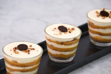 Klasik Tiramisu Tatlısı berrak bardaklarda kahve katmanları, kremalı mascarpone ve üzerine krem şanti sürülmüş kakao ve doğranmış fındıklı İtalyan tatlısının yakın görüntüsü.