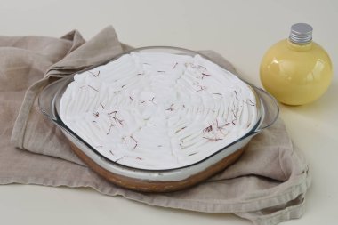 Saffron Süt Keki, beyaz kremayla kaplı cam bir fırın kabında ve kırmızı safran iplikçikleriyle süslenmiş. Süt sosu şişesi ve aromatik süt keklerinin üst görüntüsü.