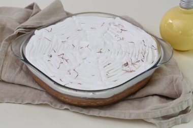 Saffron Süt Keki, beyaz kremayla kaplanmış, cam fırında pişirme kabında. Süt sosu şişesiyle süslenmiş kırmızı safran iplik. Gri peçete, sütlü keklerin üst görüntüsü.