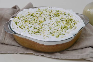 Limonlu turta şeffaf bir cam kabın içinde, üzerinde krema, parlak yeşil limon tadı ve peçeteye izole edilmiş küçük süt sosu şişesi var.