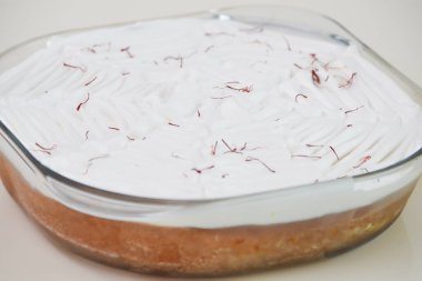 Beyaz çırpılmış kremayla kaplanmış ve aromatik sütlü keklerin yanında kırmızı safran iplikçikleriyle süslenmiş temiz bir cam fırın kabında Safran Milk Cake.