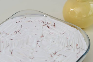 Saffron Süt Keki, beyaz kremayla kaplanmış cam bir fırın kabında ve canlı kırmızı safran iplikçikleriyle süslenmiş süt soslu, aromatik sütlü keklerin yan görüntüsü.