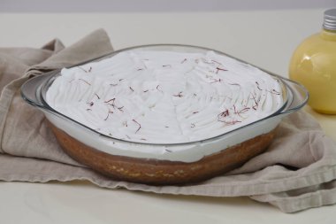 Saffron Süt Keki, beyaz kremayla kaplanmış cam bir fırın kabında ve kırmızı safran iplikçikleriyle süslenmiş. Süt sosu şişesi ve gri peçeteli aromatik sütlü kek manzarası.