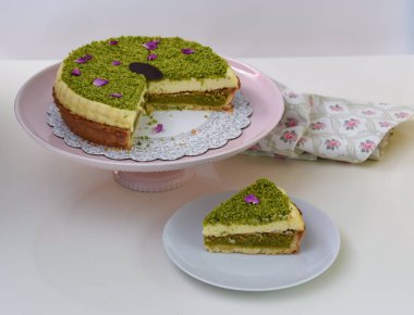Pembe pasta tezgahının üzerindeki lezzetli Pistachio Cheesecake beyaz bir tabakta sunulan dilimlenmiş fıstıklı ve mor çiçekli taçyaprağı ve peçeteli tatlı manzaralı