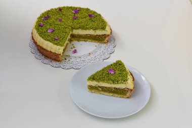 Lezzetli Pistachio Cheesecake, beyaz bir tabakta sunulan dilimlenmiş fıstık ezmesi ve mor çiçek yapraklarıyla süslenmiş zarif tatlı manzarası.