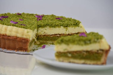 Lezzetli Pistachio Cheesecake, beyaz bir tabakta sunulan dilimlenmiş fıstıklı ve mor çiçekli taçyapraklı zarif bir tatlı.