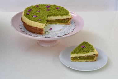 Pembe pasta tezgahının üzerindeki lezzetli Pistachio Cheesecake beyaz bir tabakta sunulan dilimlenmiş fıstıklı ve mor çiçek yapraklı klasik tatlı manzaralı