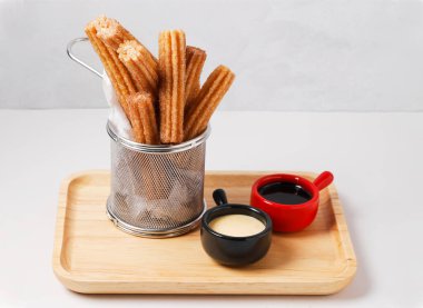 Beyaz arka planda ahşap tepsi menüde servis edilen, bol çikolatalı ve kremalı peynir soslu gevrek Altın Çurros.