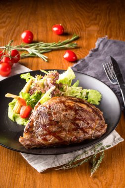 Izgara Amerikan Pirzola Bifteği, siyah tabakta sulu sığır eti taze yeşil salata, kiraz domatesleri ve yanında püre sebze.