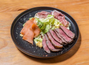Sezar Salatası Tabağı, Dilimlenmiş biftek, Tütsülenmiş Somon, ve siyah tabakta Prosciutto, ahşap tahta tahta tahta pano tarafında lüks menüler.